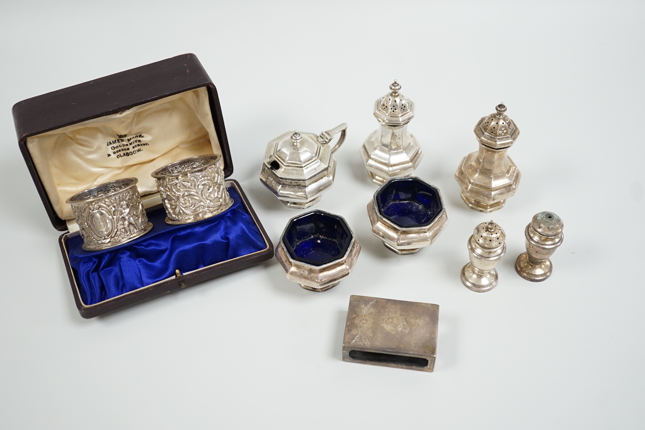 A cased pair of Edwardian embossed silver napkin rings, Birmingham, 1904, a five piece silver condiment set, Birmingham, 1926, two plated condiments and a match sleeve.